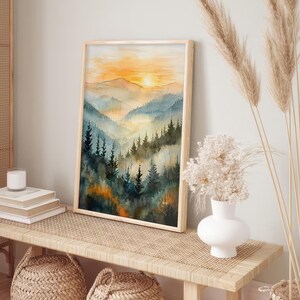 May include: A framed watercolor print of a mountain landscape. The artwork features a sunset with orange and yellow hues, and a valley filled with blue and green trees. The frame is made of light-colored wood.