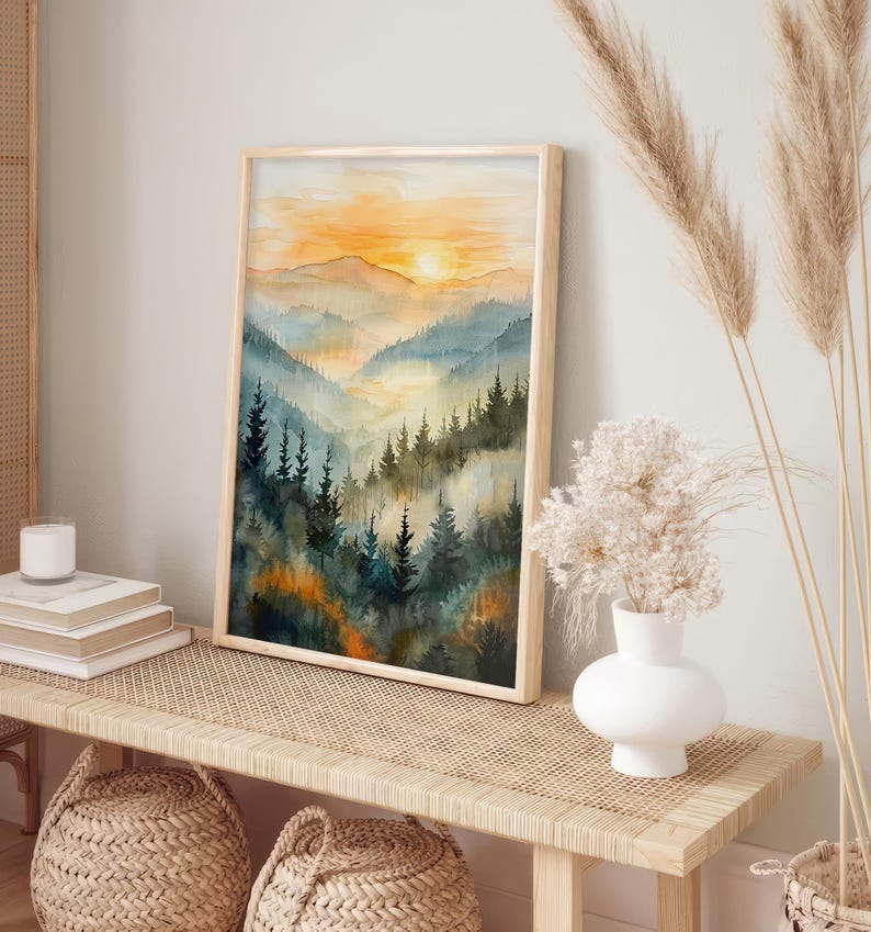 May include: A framed watercolor print of a mountain landscape. The artwork features a sunset with orange and yellow hues, and a valley filled with blue and green trees. The frame is made of light-colored wood.