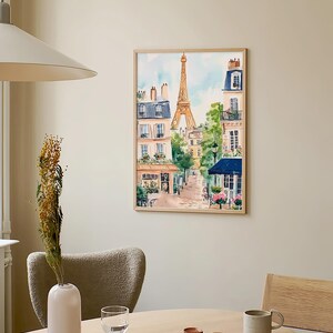 May include: A watercolour painting of a Parisian street scene with the Eiffel Tower in the background. The painting is framed in a light wood frame and is hanging on a light beige wall.