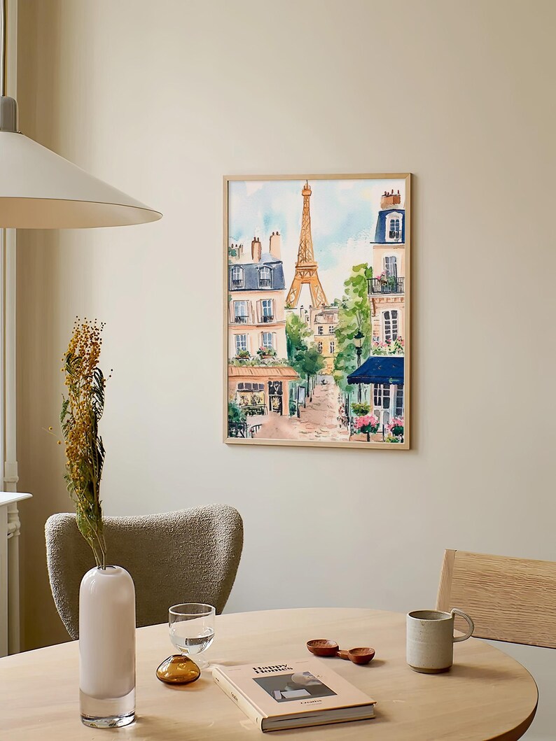 May include: A watercolour painting of a Parisian street scene with the Eiffel Tower in the background. The painting is framed in a light wood frame and is hanging on a light beige wall.