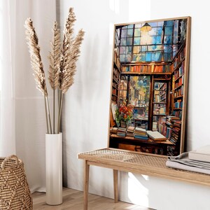 May include: A painting depicting the interior of a bookshop, featuring a large window, wooden shelves stacked with books, and a table with books and flowers. The painting is framed in a light wood frame.