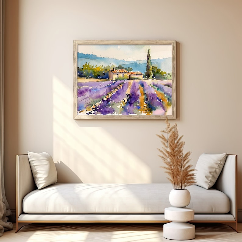 K&ouml;nnte beinhalten: Ein Aquarellbild eines Lavendelfeldes mit einem kleinen Haus in der Ferne. Das Bild ist in einem hellen Holzrahmen gerahmt und h&auml;ngt an einer hellbeigen Wand.