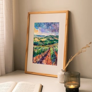 Peut inclure: Une peinture &agrave; l'aquarelle encadr&eacute;e d'un paysage de vignoble avec des collines vallonn&eacute;es, des vignes vertes et un ciel nuageux. La peinture est dans un cadre en bois et est expos&eacute;e sur une table blanche avec un livre et un bougeoir.