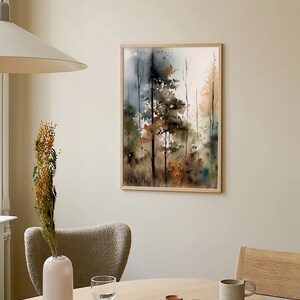 May include: A framed watercolor painting of a forest scene with trees in shades of brown, green, and blue. The painting is hanging on a light beige wall above a wooden table with a vase, a book, and a mug.