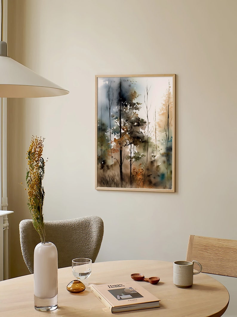 May include: A framed watercolor painting of a forest scene with trees in shades of brown, green, and blue. The painting is hanging on a light beige wall above a wooden table with a vase, a book, and a mug.