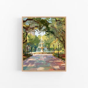 Op de afbeelding: Een impressionistisch schilderij van een parkscène met een fontein. Een pad leidt naar de fontein, omgeven door bomen en groen. Het kunstwerk is ingelijst in licht hout. Het schilderij gebruikt een palet van groen, blauw en bruin.