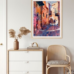 May include: Watercolor painting of a sunlit street scene in warm tones of orange, pink, and blue. The artwork is framed in a light wood frame and hangs on a neutral wall. The painting depicts buildings and figures, creating a sense of depth and atmosphere.