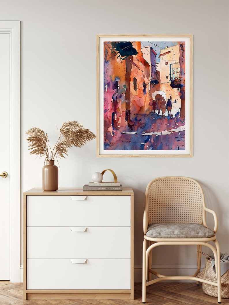May include: Watercolor painting of a sunlit street scene in warm tones of orange, pink, and blue. The artwork is framed in a light wood frame and hangs on a neutral wall. The painting depicts buildings and figures, creating a sense of depth and atmosphere.