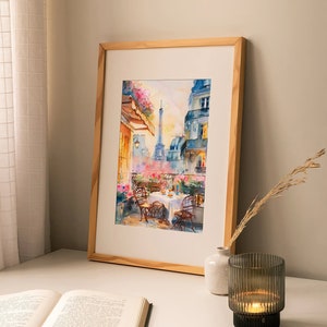 May include: A watercolor painting in a wooden frame depicts a Parisian balcony scene with a view of the Eiffel Tower. The balcony has a table set for two with flowers and a bottle of wine. The painting is on a white table with a book and a candle holder.