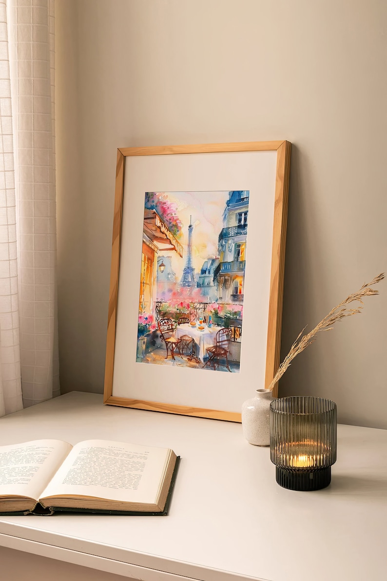 May include: A watercolor painting in a wooden frame depicts a Parisian balcony scene with a view of the Eiffel Tower. The balcony has a table set for two with flowers and a bottle of wine. The painting is on a white table with a book and a candle holder.