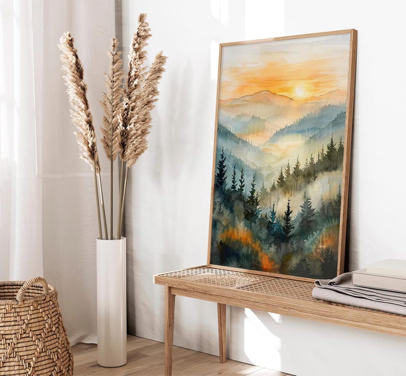 May include: A framed watercolor painting depicts a mountain landscape with a sunset. The artwork features layers of blue and green mountains, with a bright orange and yellow sky. The painting is displayed on a wooden bench.