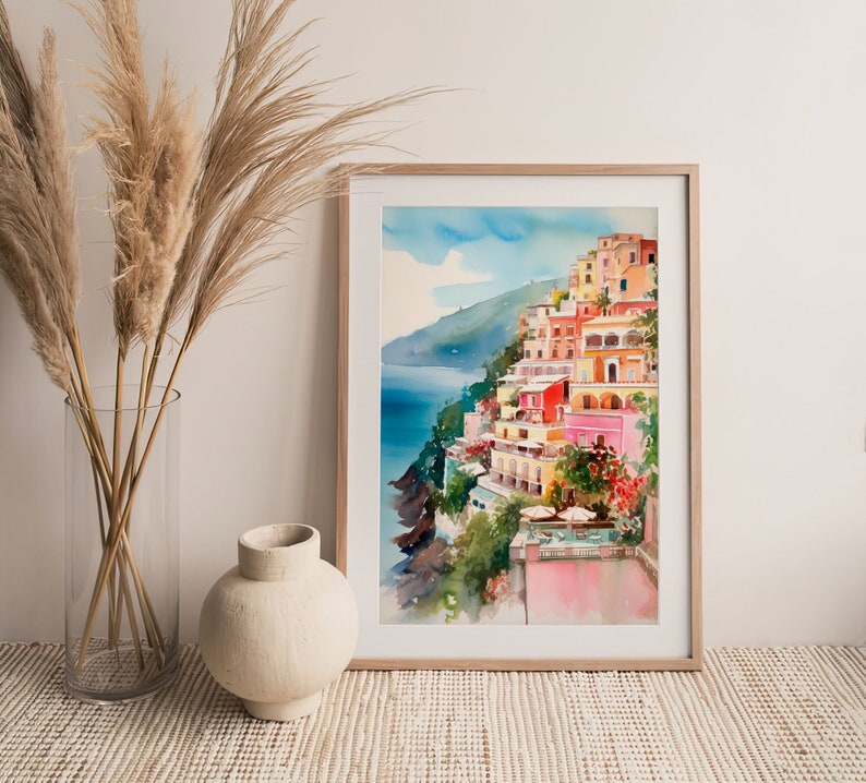 May include: A watercolour painting depicting a colourful Italian village perched on a cliff overlooking the sea. The buildings are painted in shades of pink, yellow, orange, and red. The sea is a deep blue, and the sky is a light blue with white clouds.