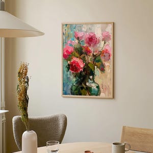 Op de afbeelding: Een ingelijst schilderij van een boeket roze rozen in een groene vaas. Het schilderij hangt aan een witte muur met een lamp erboven. Op de voorgrond staat een tafel met een witte vaas, een glas, een boek en een mok.