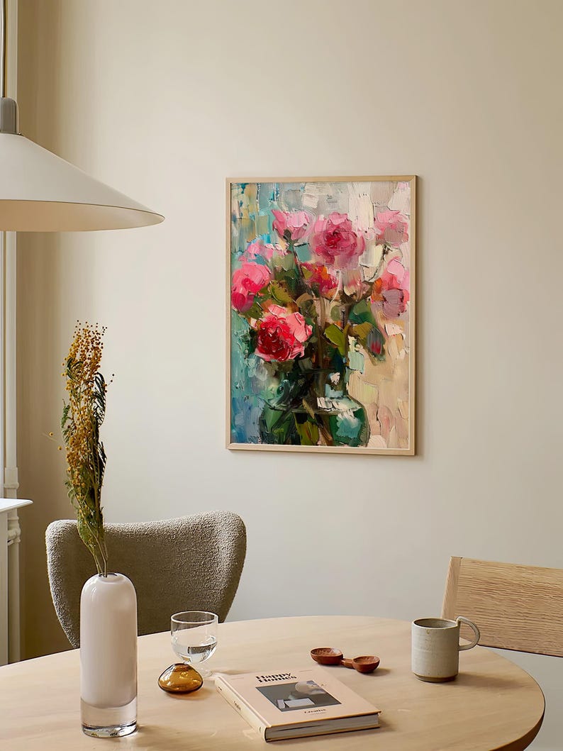 Op de afbeelding: Een ingelijst schilderij van een boeket roze rozen in een groene vaas. Het schilderij hangt aan een witte muur met een lamp erboven. Op de voorgrond staat een tafel met een witte vaas, een glas, een boek en een mok.