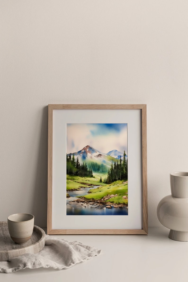 May include: A framed watercolour painting of a mountain landscape with a river flowing through a valley. The mountains are green and brown with snow-capped peaks. The river is blue and reflects the sky. The painting is set against a white background.