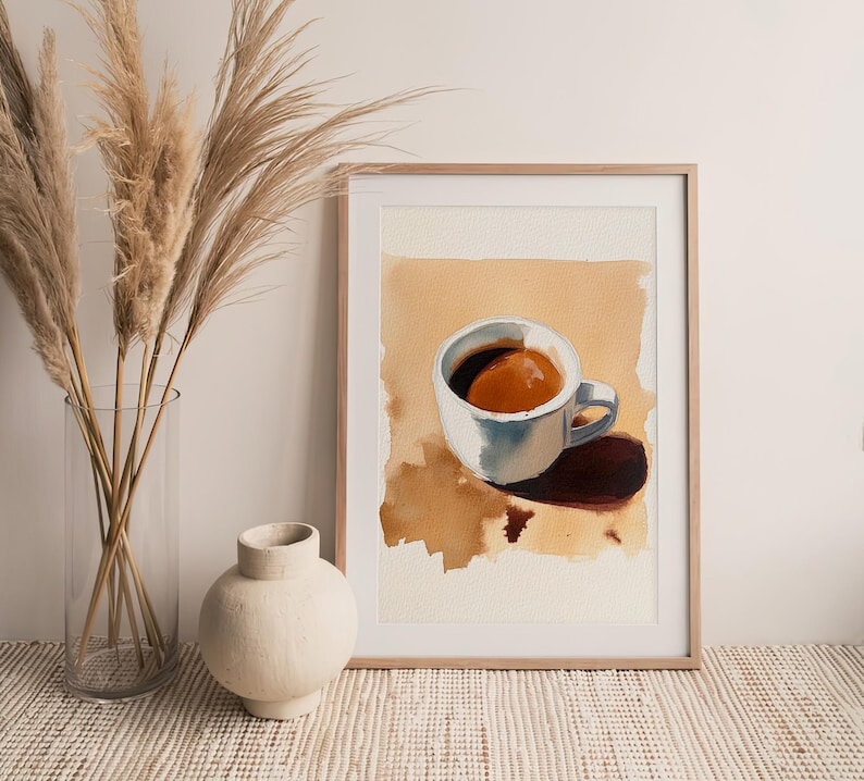 Puede incluir: Una pintura de acuarela enmarcada de una taza de caf&eacute; blanca con caf&eacute; marr&oacute;n en su interior, sentada sobre un fondo beige. La taza proyecta una sombra sobre el fondo.