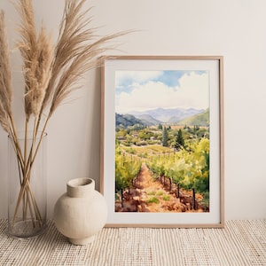 May include: A watercolor painting of a vineyard landscape with rows of grapevines leading up a hill towards a small village in the distance. The painting is framed in a light wood frame and is set against a white wall.