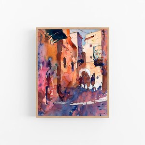 May include: Watercolor painting of a sunlit street scene with buildings in shades of orange, pink, and blue. The artwork is framed in a light wood frame. Figures are silhouetted in the archway.