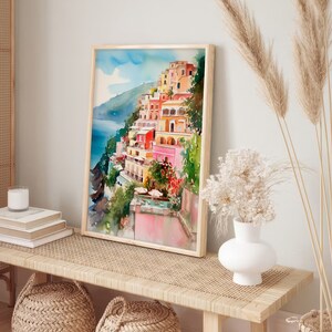 May include: A watercolour painting of a colourful Italian village on a cliffside overlooking the ocean. The painting is framed in a light wood frame and is hanging on a wall above a wooden bench with woven baskets.