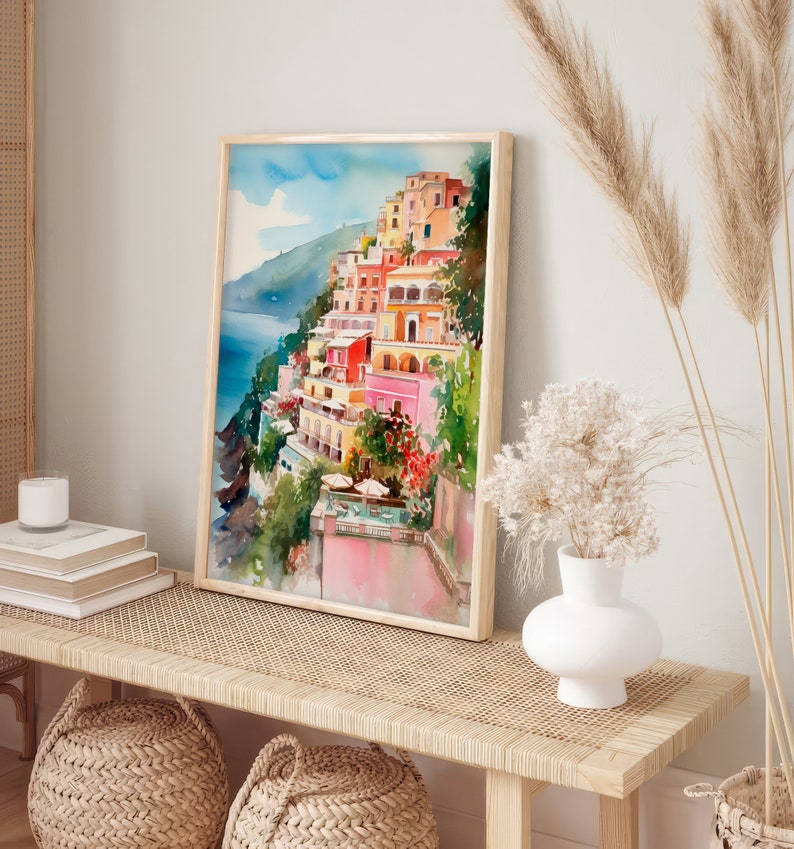May include: A watercolour painting of a colourful Italian village on a cliffside overlooking the ocean. The painting is framed in a light wood frame and is hanging on a wall above a wooden bench with woven baskets.