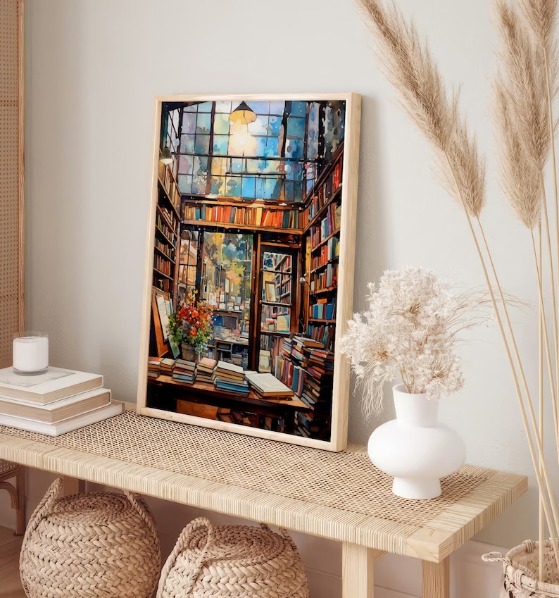 May include: A painting depicting the interior of a bookshop, featuring a large window, shelves overflowing with books, and a wooden table piled high with books. The painting is framed in a light wood frame and hangs on a white wall.