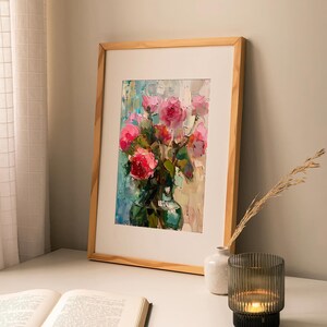 Op de afbeelding: Een ingelijst schilderij van een boeket roze rozen in een groene vaas. Het schilderij staat op een witte tafel met een boek en een kaarsenhouder.