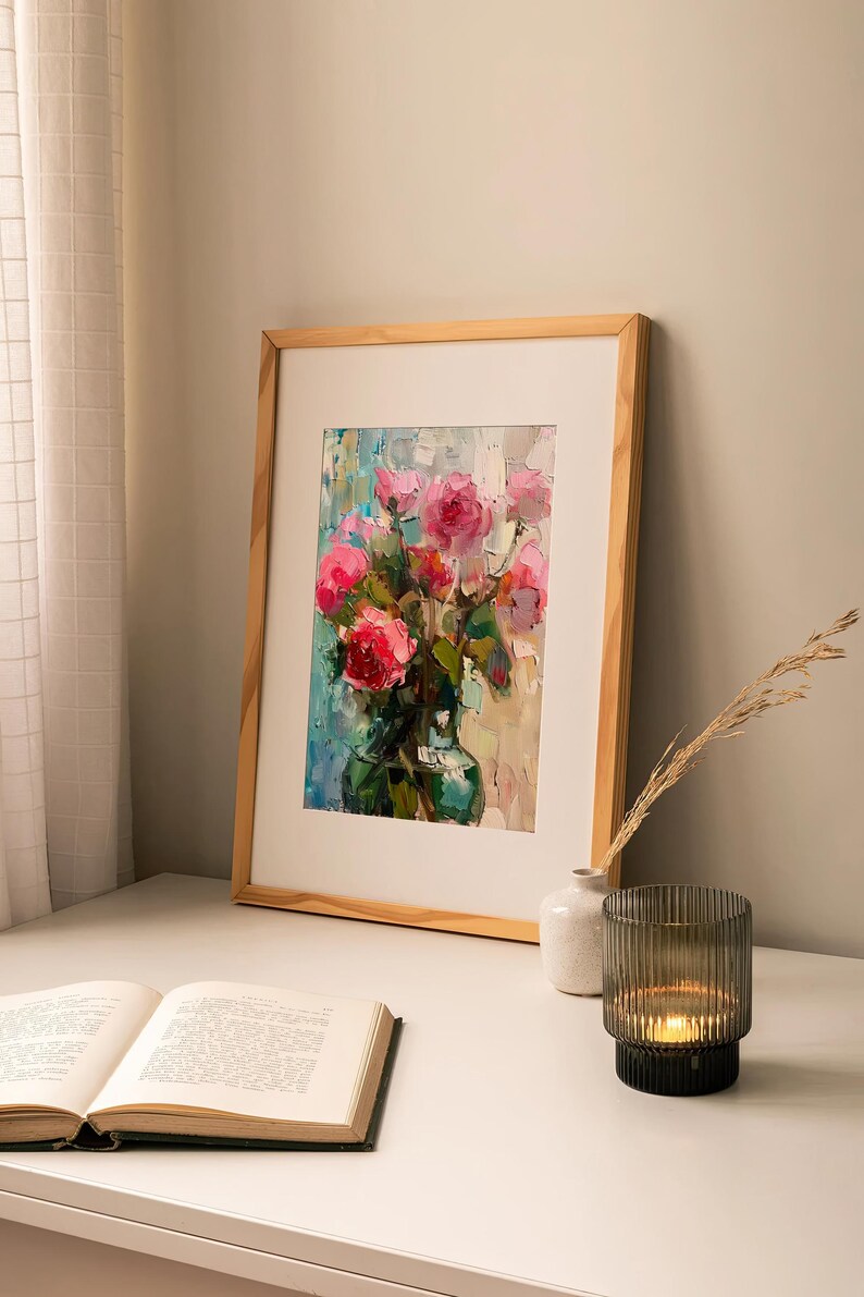 Op de afbeelding: Een ingelijst schilderij van een boeket roze rozen in een groene vaas. Het schilderij staat op een witte tafel met een boek en een kaarsenhouder.
