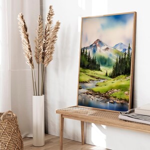 May include: A watercolour painting of a mountain landscape with a river flowing through it. The painting is framed in a light wood frame and is hanging on a white wall.