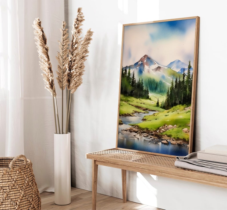 May include: A watercolour painting of a mountain landscape with a river flowing through it. The painting is framed in a light wood frame and is hanging on a white wall.