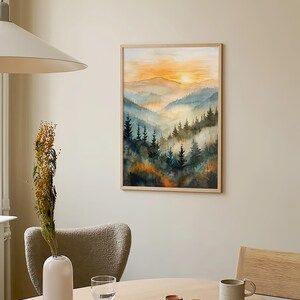 May include: A framed watercolor painting of a mountain landscape with a sunset. The artwork features layers of blue and green mountains with a vibrant orange and yellow sky. The painting is displayed on a neutral-colored wall.