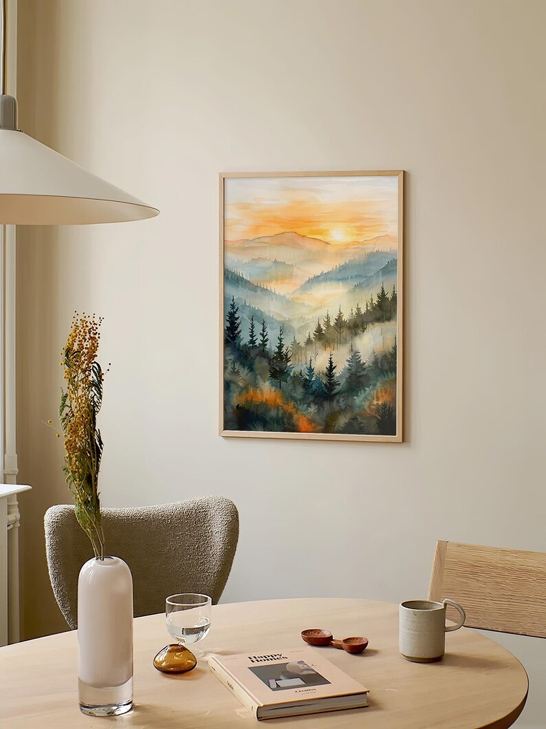 May include: A framed watercolor painting of a mountain landscape with a sunset. The artwork features layers of blue and green mountains with a vibrant orange and yellow sky. The painting is displayed on a neutral-colored wall.