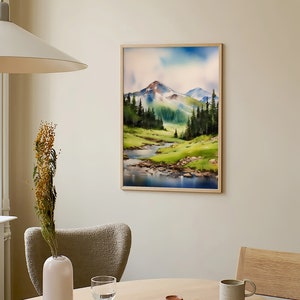 May include: A framed watercolour painting of a mountain landscape with a river flowing through a valley. The painting is in a light wood frame and is hanging on a light beige wall.