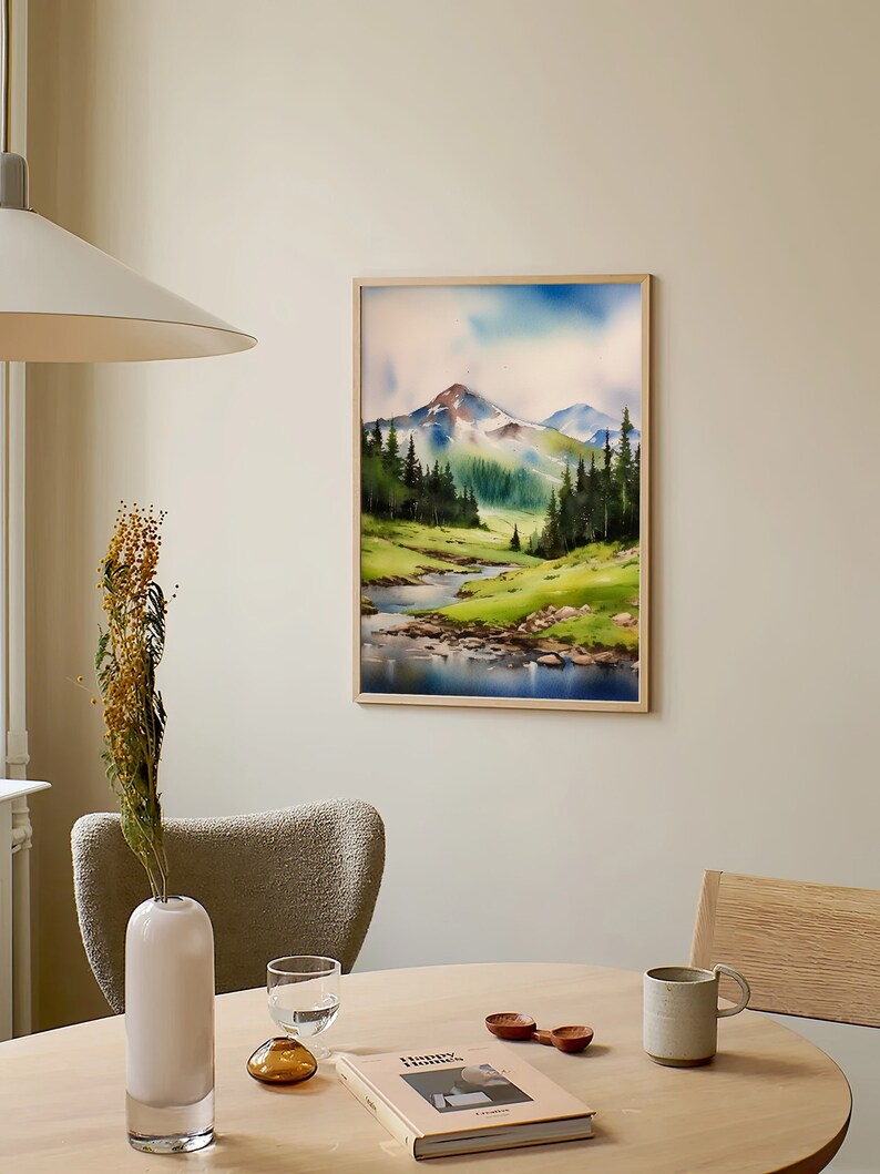 May include: A framed watercolour painting of a mountain landscape with a river flowing through a valley. The painting is in a light wood frame and is hanging on a light beige wall.