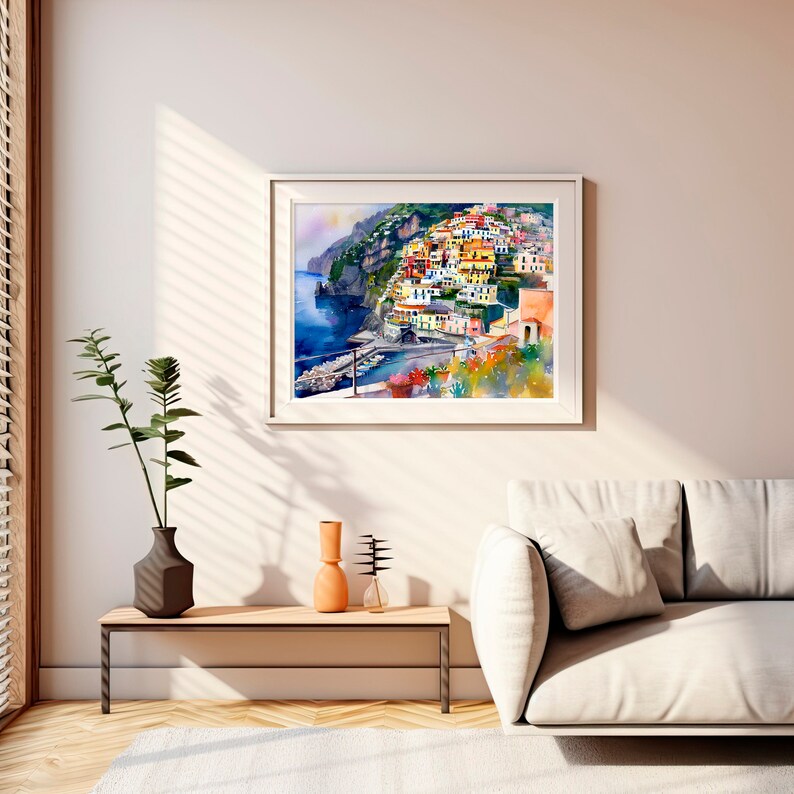 May include: A watercolour painting of a colourful Italian village nestled on a cliffside overlooking the sea. The buildings are painted in shades of yellow, orange, and red, and the water is a deep blue. The painting is framed in a white frame and hangs on a white wall.
