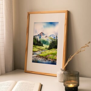 May include: A framed watercolour painting of a mountain landscape with a river flowing through a valley. The painting is in a light wood frame and is hanging on a white wall. There is a book and a candle holder on a white table in front of the painting.