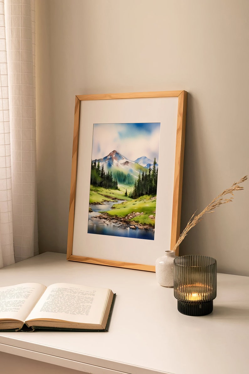 May include: A framed watercolour painting of a mountain landscape with a river flowing through a valley. The painting is in a light wood frame and is hanging on a white wall. There is a book and a candle holder on a white table in front of the painting.