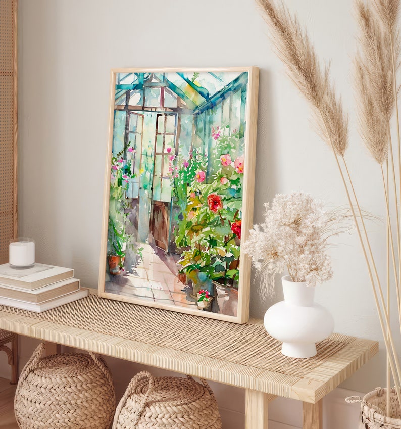 Puede incluir: Una pintura de acuarela de un invernadero lleno de vegetaci&oacute;n exuberante y flores rosas vibrantes. La pintura est&aacute; enmarcada en un marco de madera clara y cuelga de una pared blanca sobre un banco de madera.