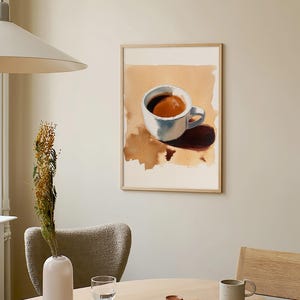 Puede incluir: Una pintura al agua enmarcada de una taza de caf&eacute; blanca con caf&eacute; marr&oacute;n en su interior. La pintura est&aacute; colgada en una pared de color beige claro sobre una mesa de madera con un jarr&oacute;n, un vaso, un libro y una taza de cer&aacute;mica.