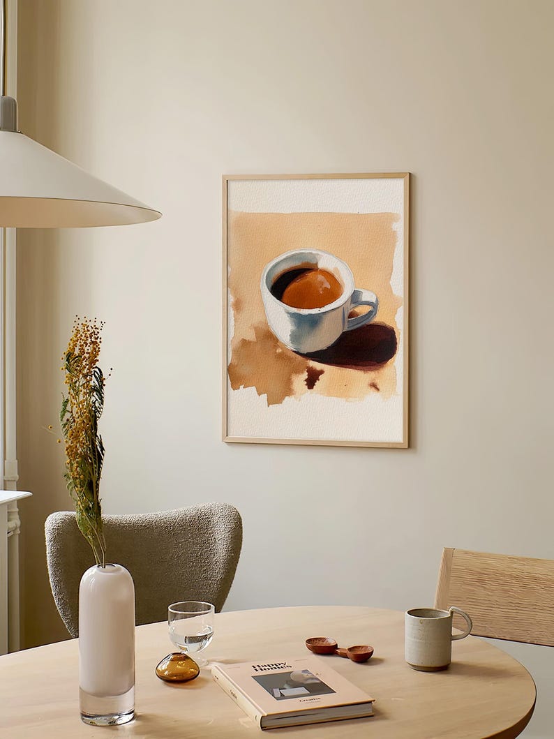 Puede incluir: Una pintura al agua enmarcada de una taza de caf&eacute; blanca con caf&eacute; marr&oacute;n en su interior. La pintura est&aacute; colgada en una pared de color beige claro sobre una mesa de madera con un jarr&oacute;n, un vaso, un libro y una taza de cer&aacute;mica.