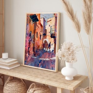 May include: Watercolor painting of a street scene in warm tones, framed in light wood. The artwork depicts buildings and figures with shades of orange, purple, and blue. The painting is displayed on a woven bench with decorative items.