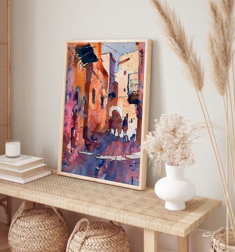 May include: Watercolor painting of a street scene in warm tones, framed in light wood. The artwork depicts buildings and figures with shades of orange, purple, and blue. The painting is displayed on a woven bench with decorative items.
