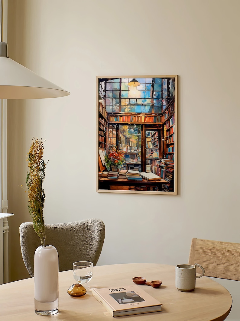 May include: A framed painting depicting the interior of a bookshop, featuring a large window, wooden shelves stacked with books, and a table with books and a vase of flowers. The painting is in a light wood frame and is hanging on a beige wall.