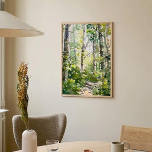 May include: A framed watercolor painting of a forest scene with tall trees and green foliage. The painting is hanging on a light beige wall above a wooden table with a vase, a glass, a book, and a mug.