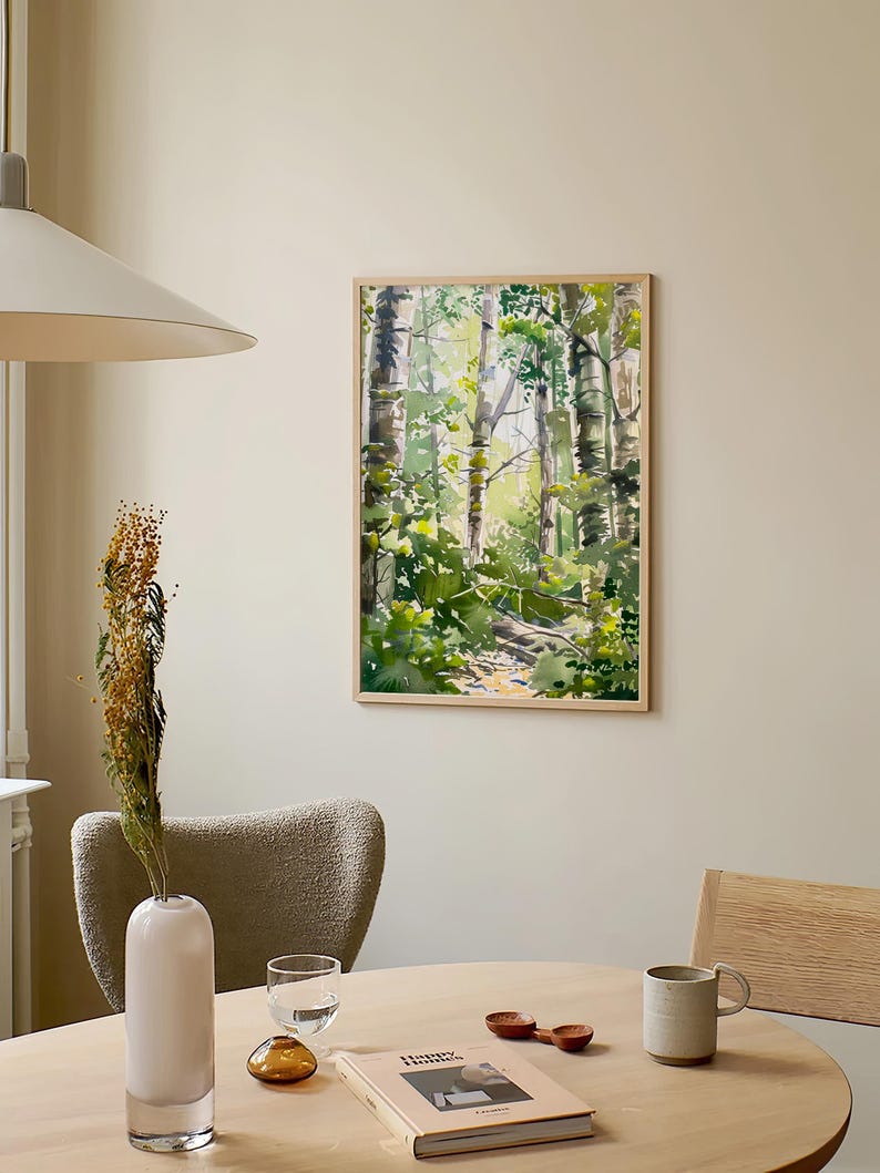 May include: A framed watercolor painting of a forest scene with tall trees and green foliage. The painting is hanging on a light beige wall above a wooden table with a vase, a glass, a book, and a mug.