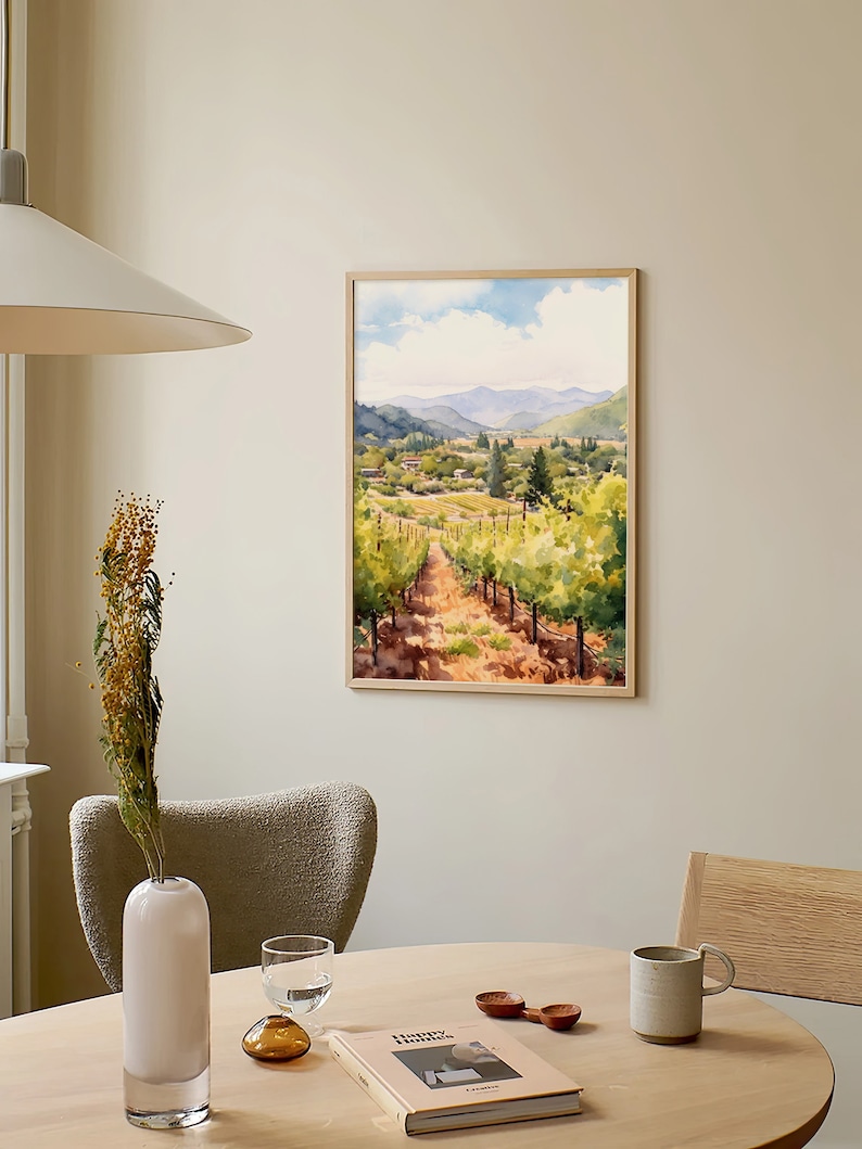 May include: A framed watercolor painting of a vineyard landscape with rolling hills, green vines, and a dirt path leading through the center. The painting is in a light wood frame and hangs on a beige wall.
