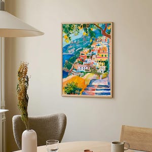 May include: A framed watercolour painting shows a vibrant coastal village on a hillside. The artwork uses bright colours like orange, yellow, blue, and green, with a tree in the upper left. The painting is displayed on a neutral wall.