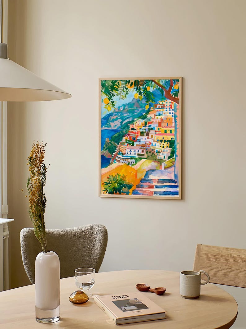 May include: A framed watercolour painting shows a vibrant coastal village on a hillside. The artwork uses bright colours like orange, yellow, blue, and green, with a tree in the upper left. The painting is displayed on a neutral wall.