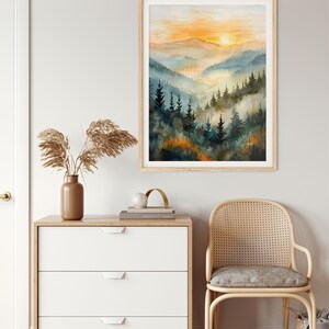 May include: A framed watercolor painting of a mountain landscape with a sunset. The artwork features layers of blue and green trees and hills, with an orange and yellow sky. The frame is light wood.