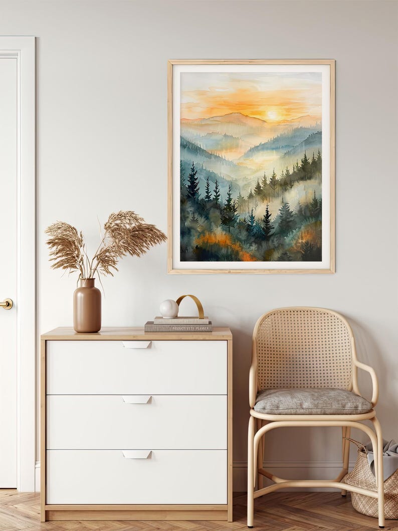 May include: A framed watercolor painting of a mountain landscape with a sunset. The artwork features layers of blue and green trees and hills, with an orange and yellow sky. The frame is light wood.
