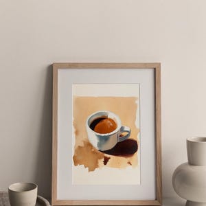 Puede incluir: Una pintura de acuarela enmarcada de una taza de caf&eacute; blanca con un l&iacute;quido marr&oacute;n en su interior. La taza est&aacute; sentada sobre una superficie marr&oacute;n claro con una sombra debajo. La pintura est&aacute; en un marco de madera claro.